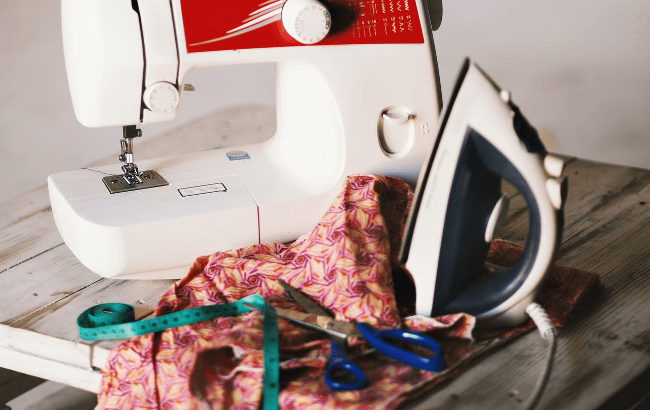 Wooden table with sewing machine, scissors, measuring tape, an iron and patterned material lying on it